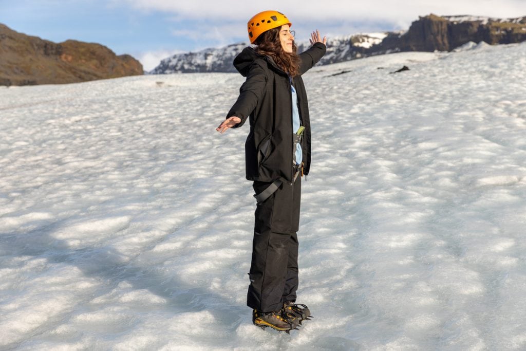 A woman stands on a glacier with her arms outstretched, wearing a helmet, crampons, and cold-weather gear. Snow-covered mountains rise in the background under a partly cloudy sky, highlighting the vast icy terrain.