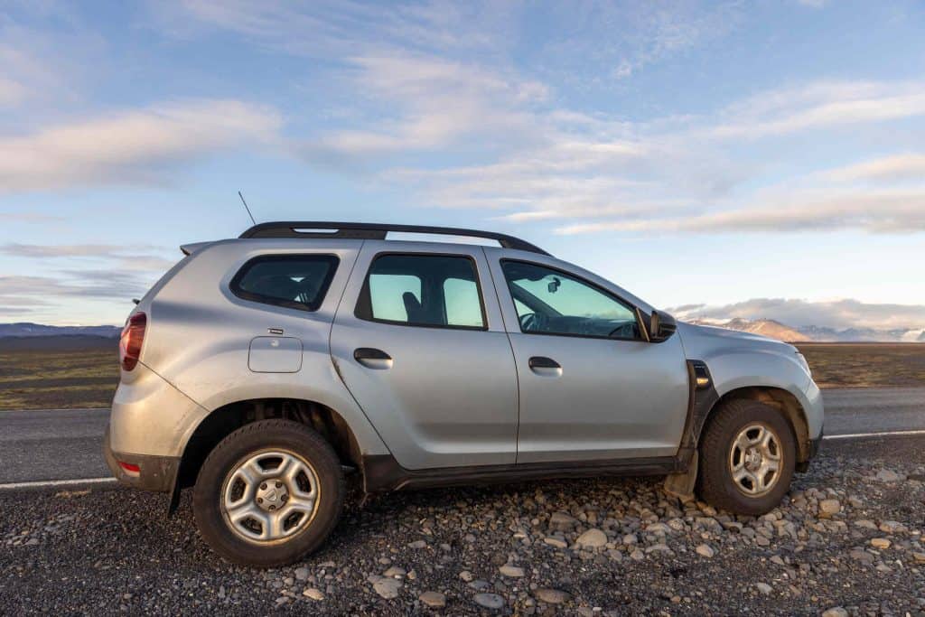 A silver SUV with off-road tires is parked on a gravel shoulder beside an empty road, with a wide open landscape and distant mountains under a partly cloudy sky. The lighting suggests early morning or late afternoon, highlighting the rugged setting.