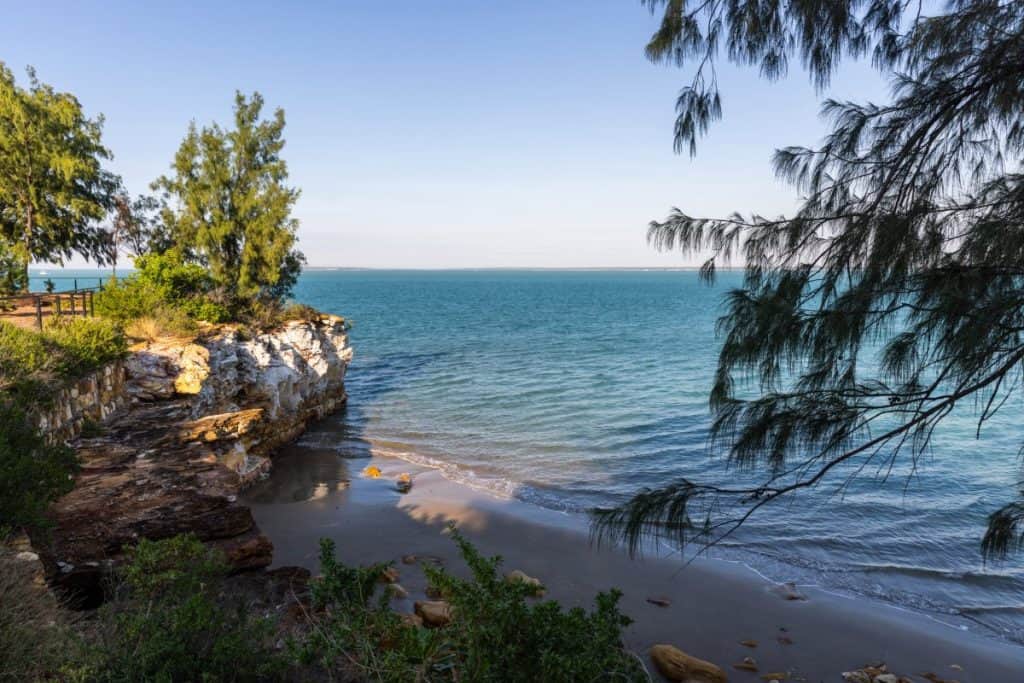A serene coastal scene with turquoise waves gently lapping against a sandy beach and rocky cliffs, framed by leafy trees under a clear blue sky.