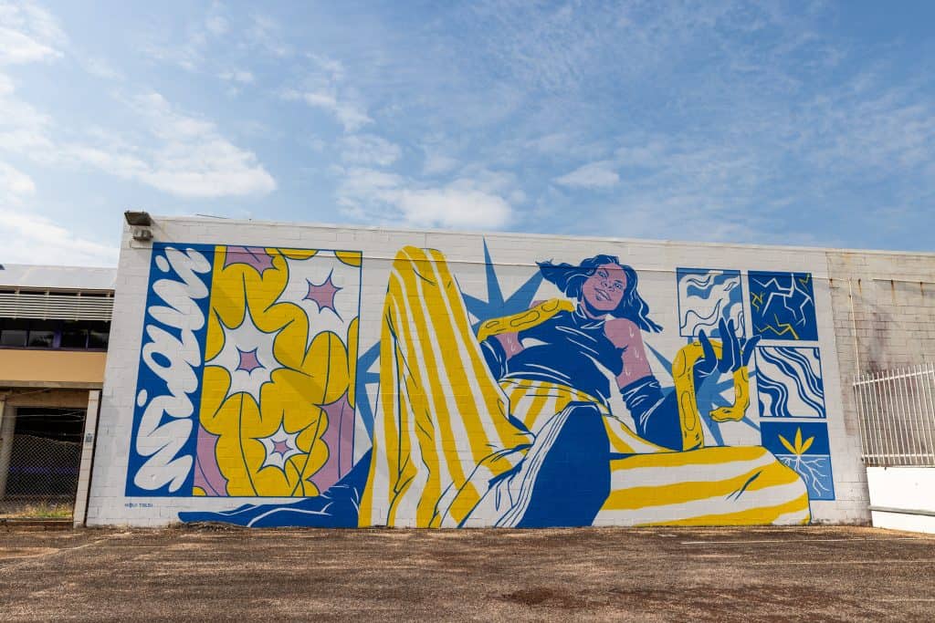 A vibrant street mural features a relaxed woman in blue and yellow clothing surrounded by bold floral and graphic elements, painted on the side of a white building under a bright blue sky.