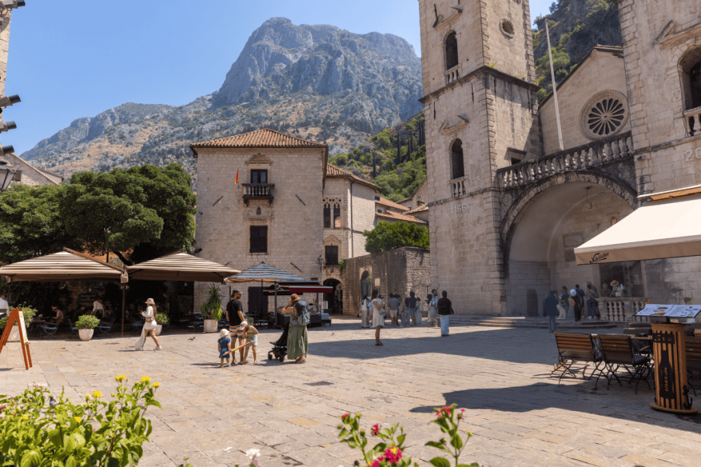 A sunny stone plaza with people strolling and relaxing near café umbrellas, surrounded by historic buildings and a grand cathedral with twin bell towers. Towering rocky mountains rise dramatically in the background, adding to the old-world charm of the scene.