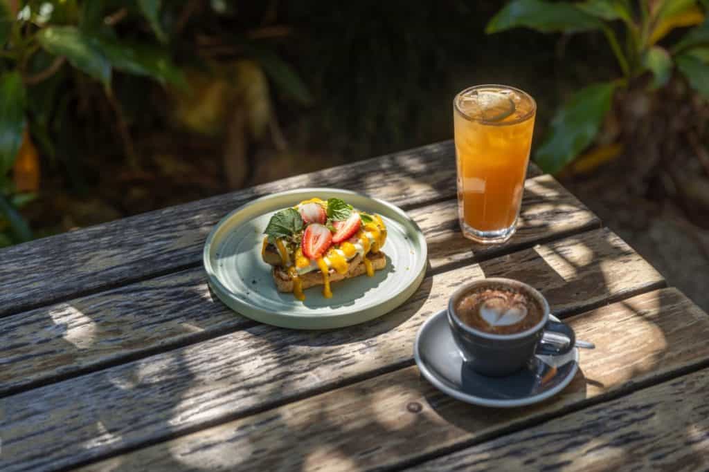 A wooden picnic table holds a plate of waffles topped with fresh strawberries and herbs, a cappuccino, and a glass of iced tea, set in a shaded garden cafe.