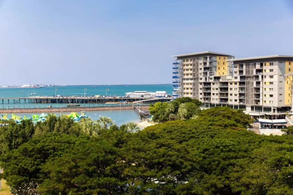 A view of a coastal city features modern high-rise apartments, a wooden pier, and a waterfront recreation area with inflatable structures on the water, all backed by a calm blue sea.
