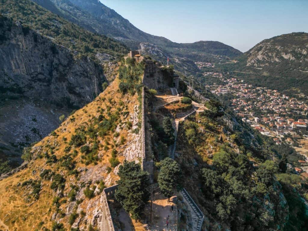 Aerial view of the San Giovanni Fortress in Kotor, Montenegro, perched on a steep hill with ancient stone walls winding up the rugged terrain. The fortress overlooks a valley with a densely packed town nestled between dramatic green mountains.