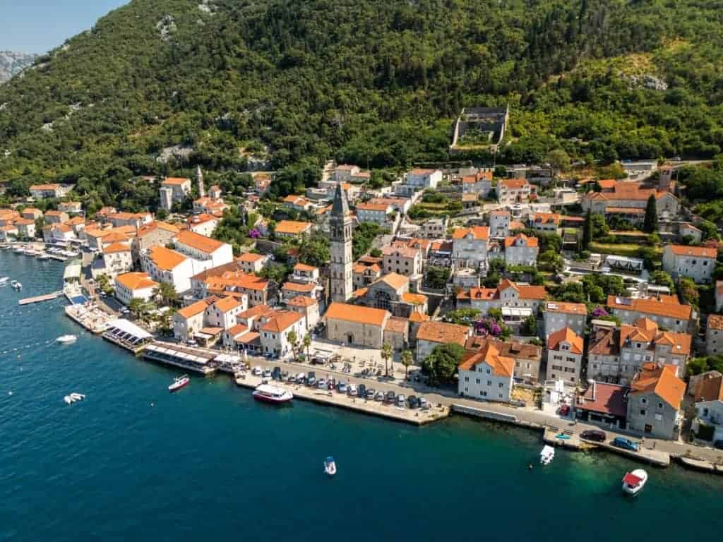 Aerial view of a coastal town with terracotta rooftops lining the edge of a clear blue bay, nestled against a lush green hillside. A tall stone bell tower rises from the center, surrounded by narrow streets, small boats, and waterfront cafes.