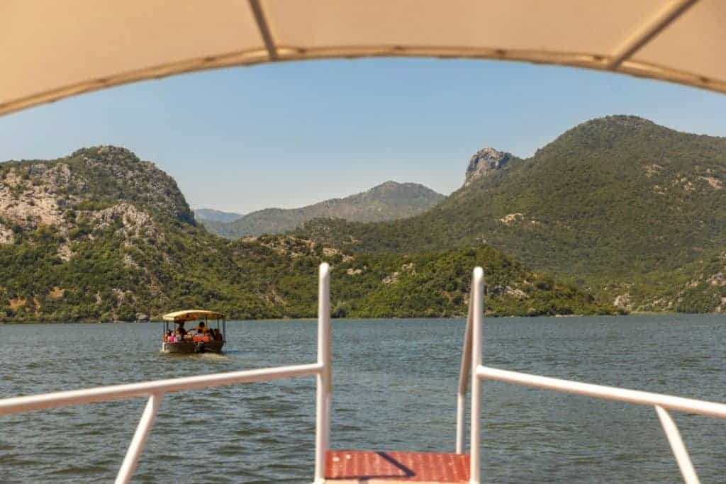 View from a boat heading across a calm lake, framed by white railings and canopy, with another boat carrying passengers visible ahead. Lush green mountains and rocky hills surround the water under a clear blue sky.