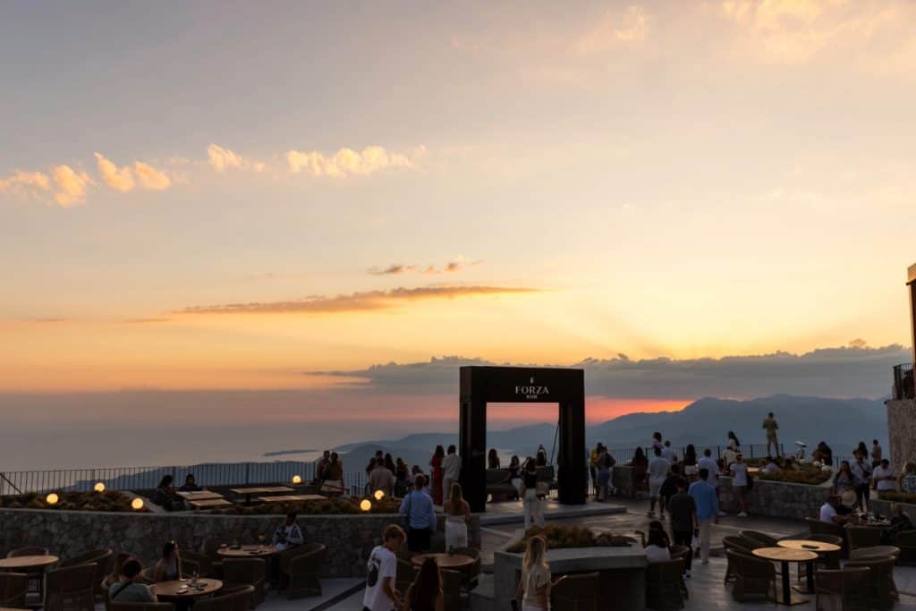 Scenic sunset at a mountaintop venue with people gathered around stone terraces and outdoor seating at “Forza,” overlooking a panoramic view of the sea and distant mountains.