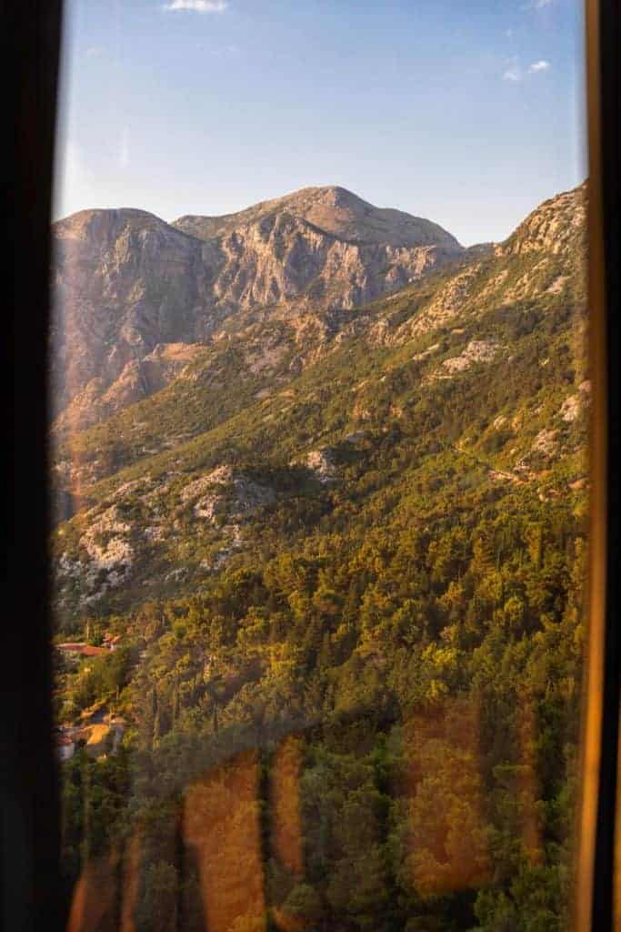 Scenic view of rugged mountains and dense forest seen through a window, with warm sunlight casting reflections and highlighting the texture of the landscape.