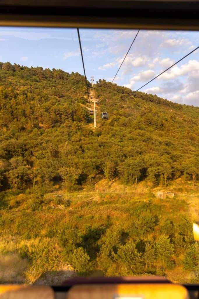View through a window of a cable car moving over a lush green mountainside during golden hour, with cables and support towers visible stretching across the frame.