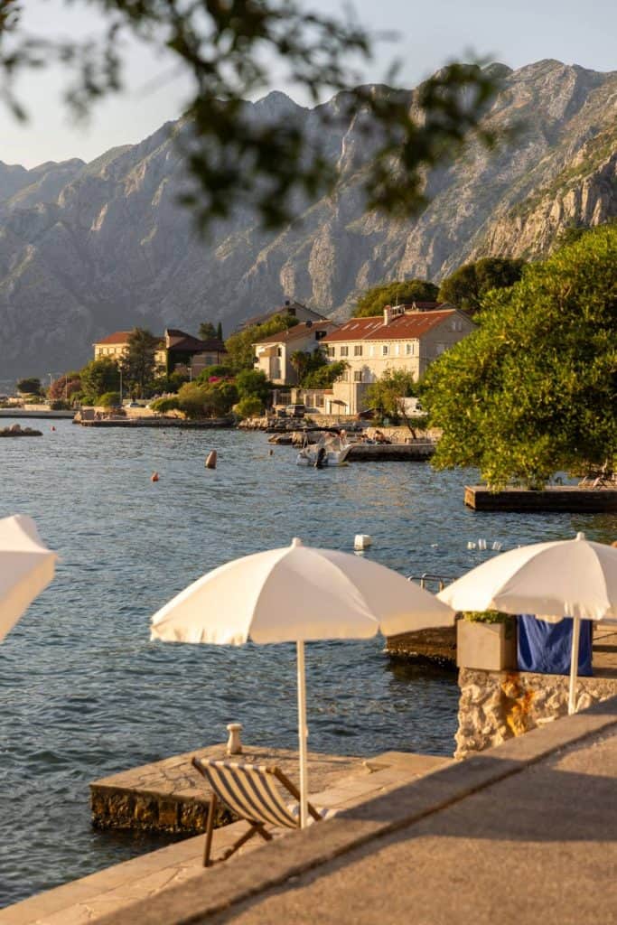 White beach umbrellas and a wooden lounge chair sit on a stone pier overlooking a calm bay with coastal houses and dramatic mountain cliffs in the background. The warm evening light casts a golden glow over the scene.