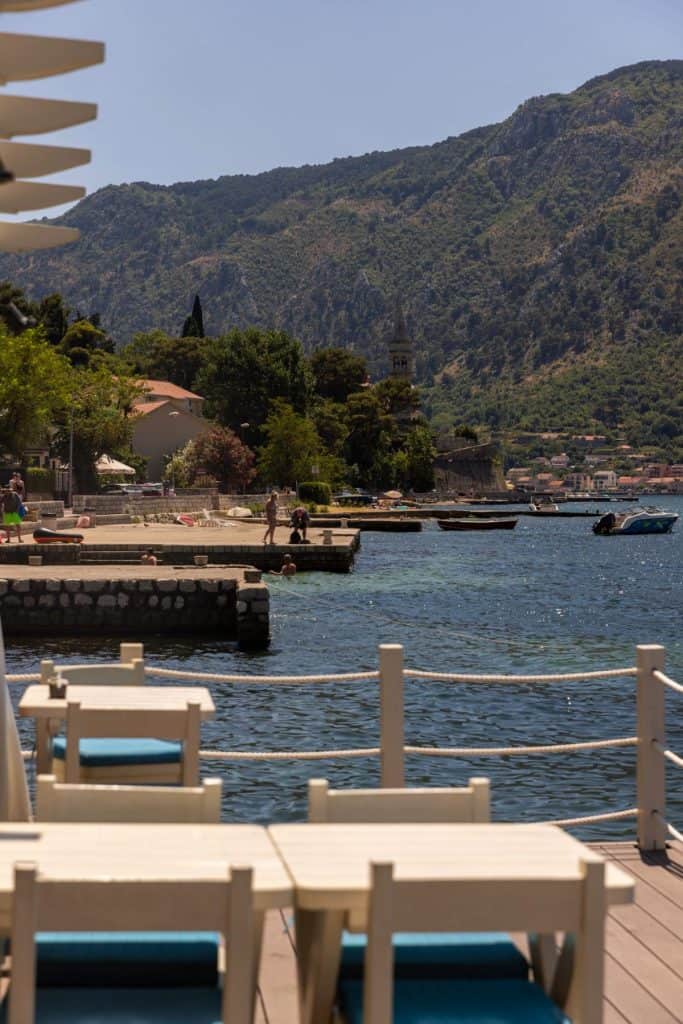 Outdoor dining tables with white chairs line a waterfront deck, facing a scenic bay surrounded by lush green mountains and seaside homes. The midday sun brightens the peaceful atmosphere and clear blue water.