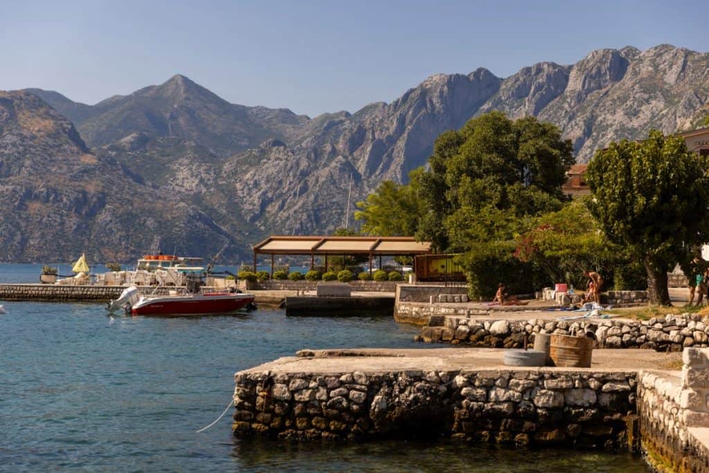 Stone piers curve along the edge of a calm bay, where people sunbathe near the water and a red motorboat is docked. In the background, rugged gray mountains rise sharply, adding dramatic contrast to the coastal village and lush greenery.