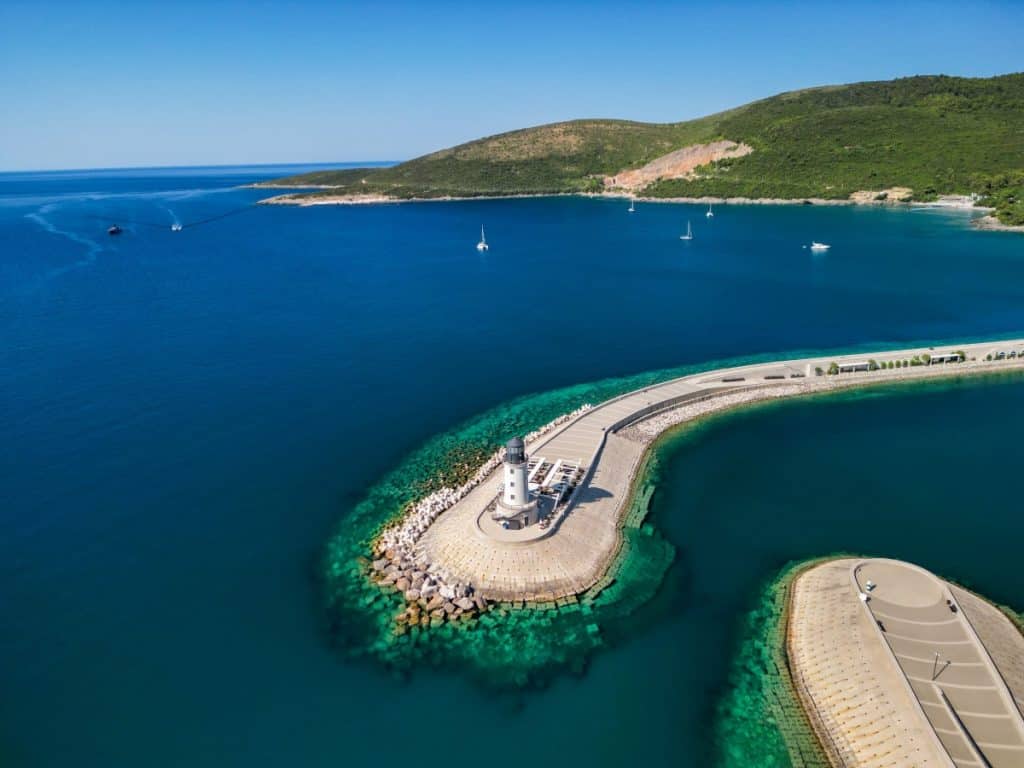 Aerial view of a white lighthouse perched on a curved stone pier surrounded by turquoise waters, with a few sailboats scattered across the deep blue bay. The lush green hillside in the background contrasts with the sleek, modern harbor design.
