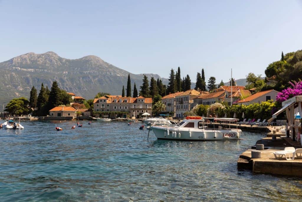 A small white boat named "Loro" floats near a peaceful waterfront village, with stone buildings and terracotta rooftops backed by tall cypress trees. In the background, mountains rise under a clear blue sky, while the foreground shows crystal-clear water and a sun-soaked stone dock with lounge chairs.