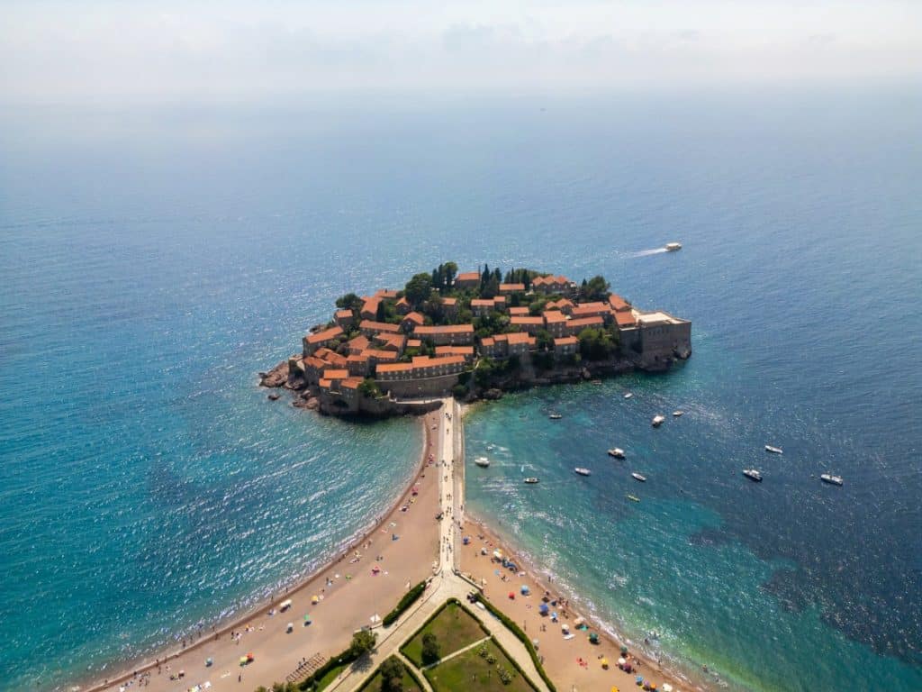 Aerial view of Sveti Stefan, a small fortified island village in Montenegro, connected to the mainland by a narrow isthmus with a sandy beach on both sides. The island is dotted with red-roofed buildings surrounded by turquoise waters and scattered boats, with a speedboat cruising nearby.