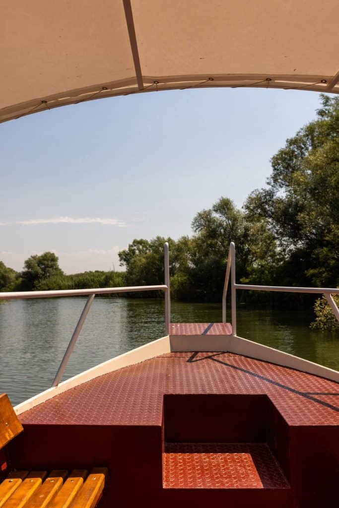 View from a boat's shaded deck looking out toward a calm river surrounded by lush green trees under a clear blue sky. The red metal floor and railing of the boat are visible in the foreground.
