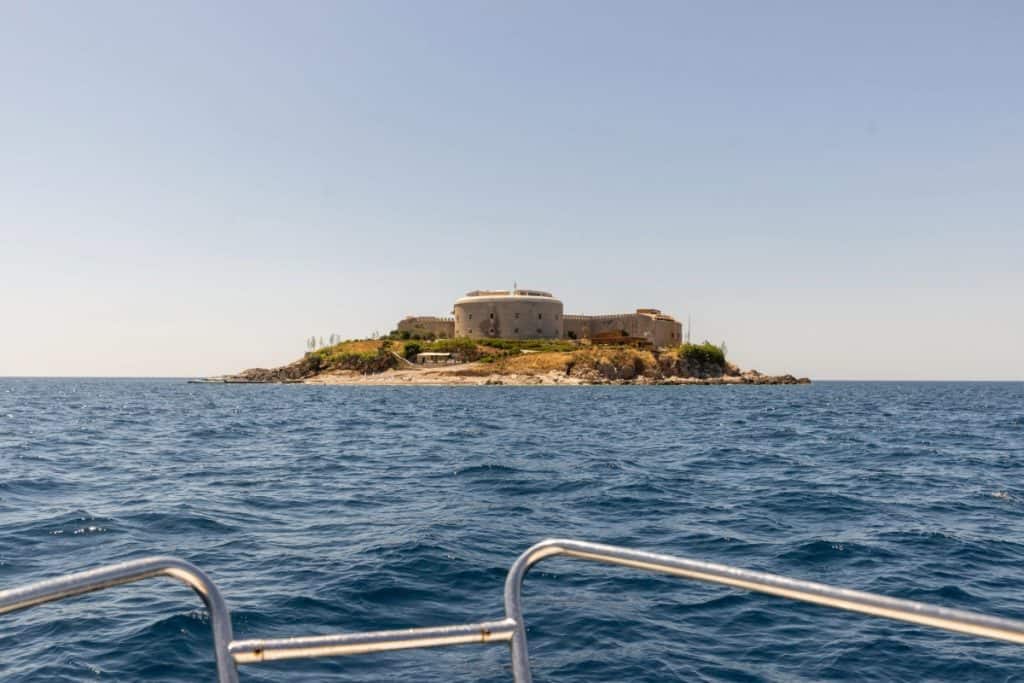 View from a boat approaching a small rocky island with a historic stone fortress in the center, surrounded by deep blue sea under a cloudless sky. The boat’s metal railing frames the foreground of the image.