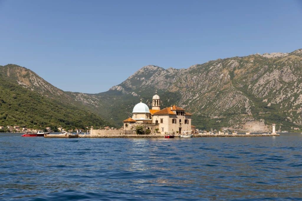 A small island with a historic church featuring a white dome and orange-tiled roofs sits in the Bay of Kotor, Montenegro, surrounded by deep blue water and backed by steep, green mountains. Several boats are docked nearby, emphasizing the island's remote and scenic location.
