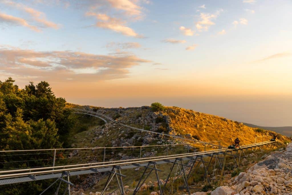A single rider is seen on an alpine coaster track winding through rocky hills and grassy slopes, surrounded by trees on the left and golden-hour sunlight casting a warm glow over the scene. The sky is streaked with soft clouds, enhancing the serene mountain landscape.