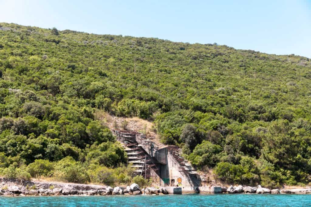 Image shows a hidden, deteriorating naval tunnel entrance built into a lush, green hillside along the coast, with concrete structures and rusted metal framing partially obscured by vegetation. The tunnel sits right at the edge of the turquoise water, hinting at its former use as a submarine or military vessel shelter.