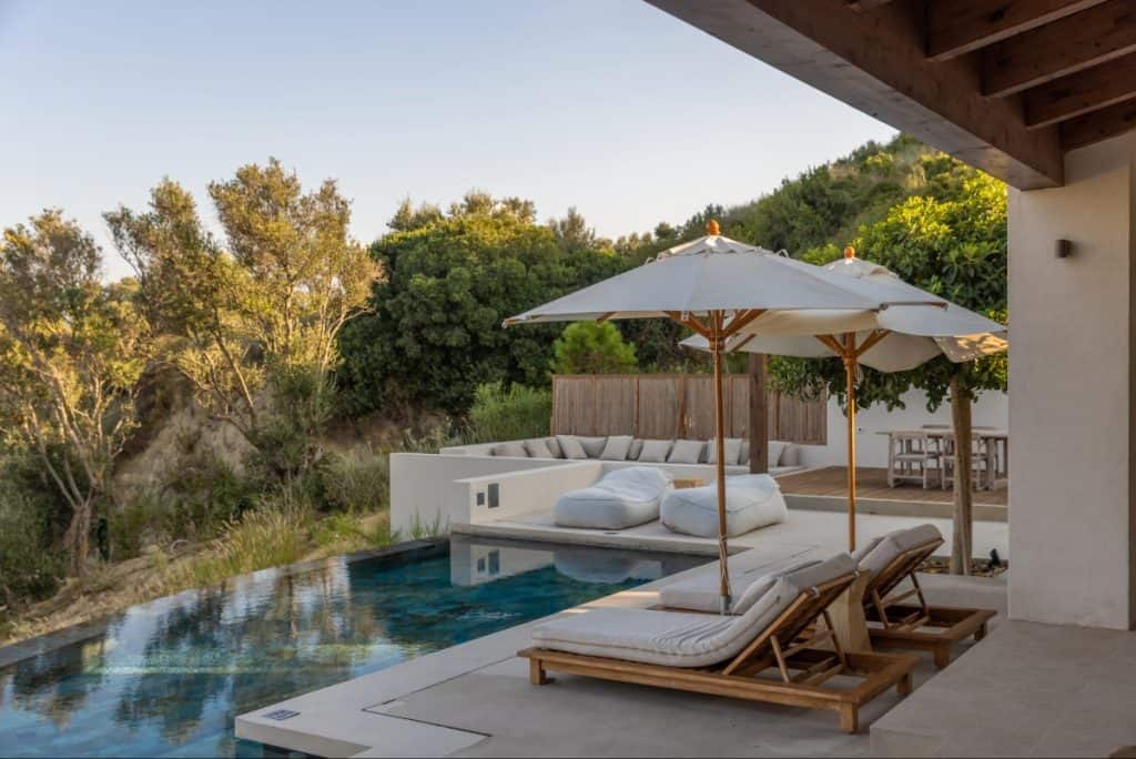 Serene outdoor area of a luxury villa featuring cushioned lounge chairs and white umbrellas beside an infinity pool. The scene is surrounded by lush greenery and modern outdoor seating, evoking a peaceful, upscale retreat.
