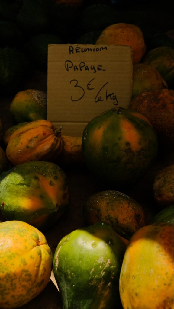 Pile of green and orange papayas displayed at a market stall with a handwritten cardboard sign reading “Réunion Papaye 3€/kg” in the center.