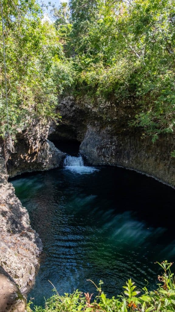 Small waterfall flowing into a circular, dark pool beneath an overhanging rock ledge, with trees and greenery above and around the canyon.