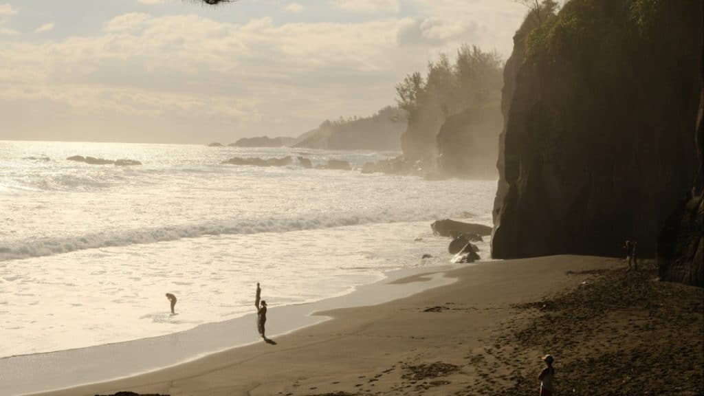 Late afternoon view of a black sand beach bordered by tall cliffs and misty waves, with a few people playing at the shoreline under soft golden light.