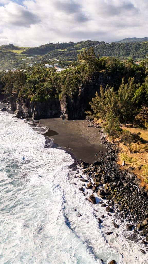 Aerial view of a rugged black sand beach with crashing white waves, surrounded by steep cliffs and dense tropical greenery on Réunion Island. Small houses are visible on the lush hillside in the background.
