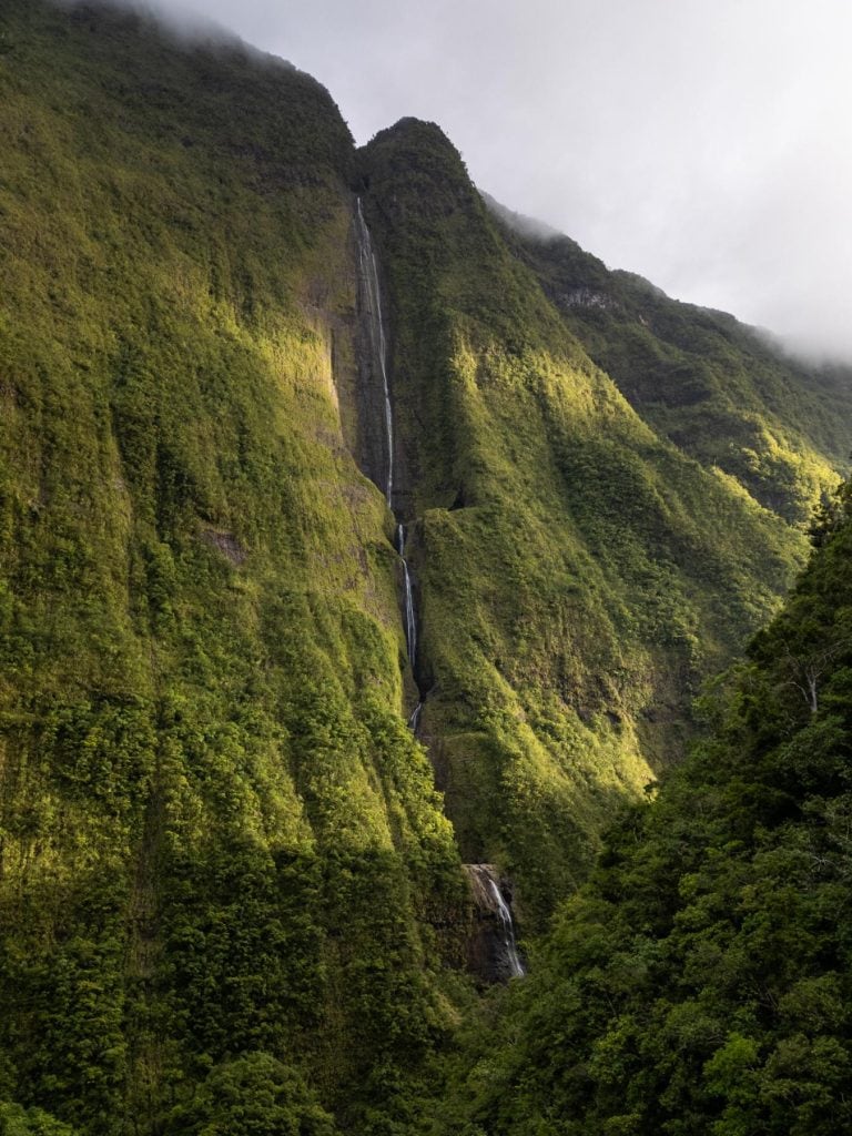Tall, narrow waterfall cascading down a steep green mountain face partially lit by sunlight, with the peak disappearing into misty clouds.