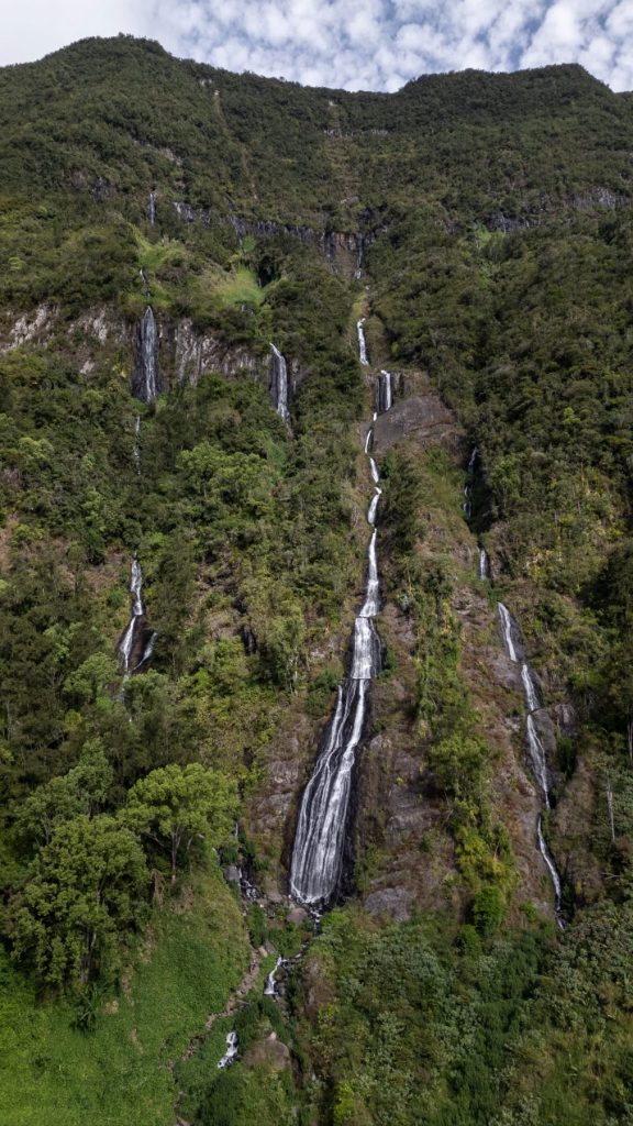 Tall, narrow waterfalls stream down a high, forested cliff in multiple tiers, surrounded by rich greenery and framed by treetops at the base.