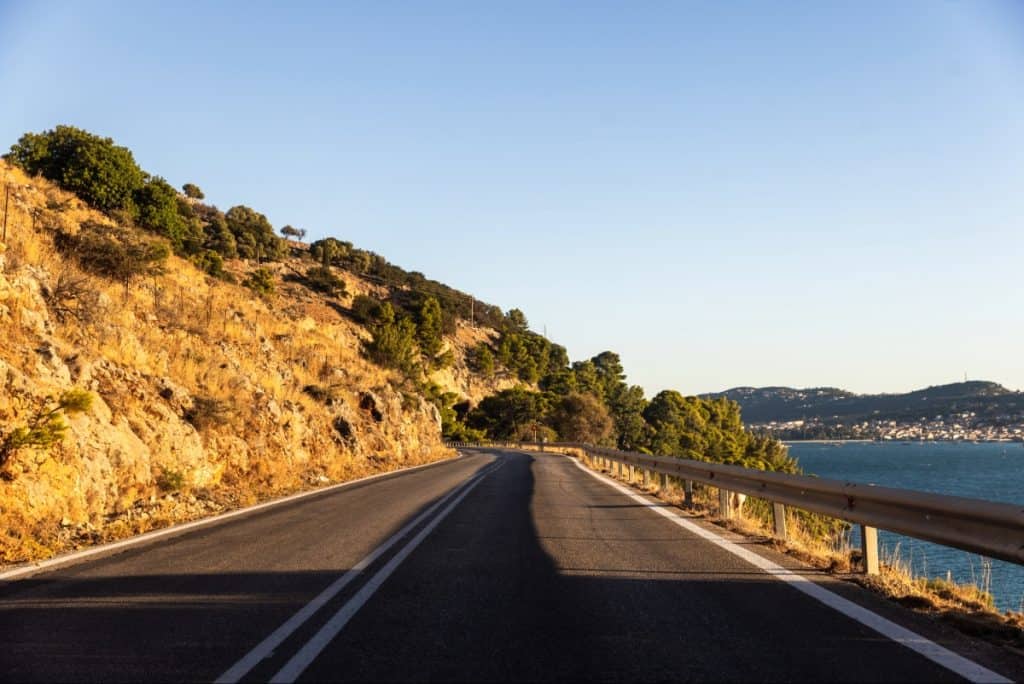 A coastal road curves gently along a sunlit cliffside, bordered by a dry, rocky slope with scattered trees on the left and a wooden guardrail overlooking the sea on the right. The scene captures the late afternoon light casting long shadows, with a distant town visible across the water.