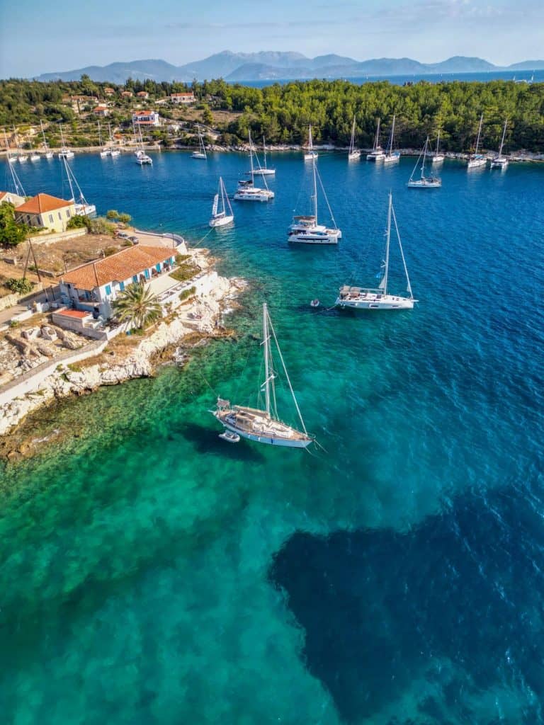 Aerial view of a serene coastal village with clear turquoise waters, several anchored sailboats, and charming stone houses with terracotta roofs nestled among lush greenery. Mountains rise in the distant background under a clear blue sky.