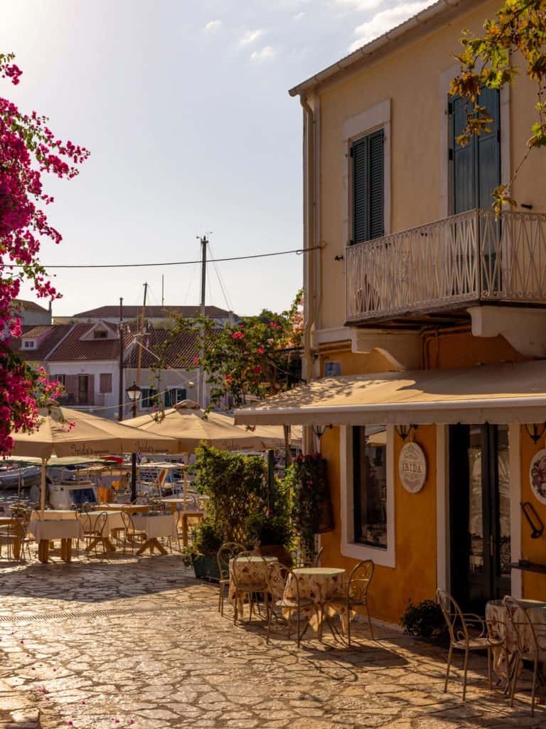 Cobblestone street lined with umbrella-shaded café tables and pastel-colored buildings in a quiet coastal village. The golden hour light bathes the scene, highlighting flowering vines and calm marina vibes.