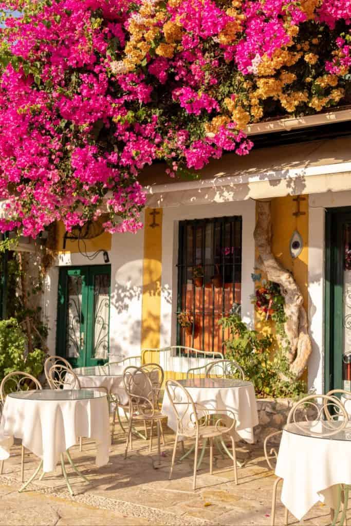 Outdoor seating area of a quaint Mediterranean café, with round tables draped in white cloths beneath vibrant pink and yellow bougainvillea. The sunlit building features green shutters, potted plants, and a rustic charm.