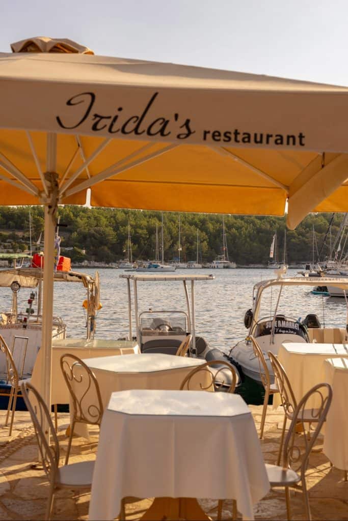 Outdoor dining tables under a large yellow umbrella labeled "Irida’s restaurant," set on a stone terrace by a marina. Small boats and sailboats float in the background, framed by tree-covered hills and warm afternoon light.
