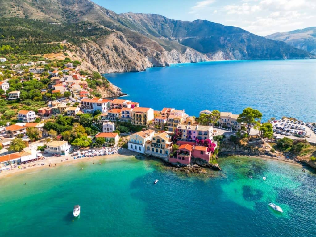 Aerial view of a colorful coastal village with pastel buildings clustered along the shoreline, nestled between clear blue waters and rugged mountain cliffs. Boats float in the turquoise sea, and a small sandy beach is visible near the village center.