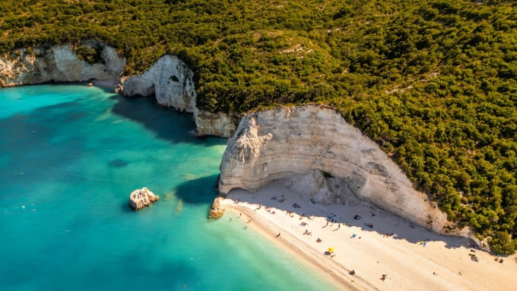 Aerial view of a secluded beach with turquoise waters, white pebble shoreline, and dramatic limestone cliffs covered in dense greenery. Sunbathers and umbrellas are scattered along the beach, adding scale to the natural landscape.
