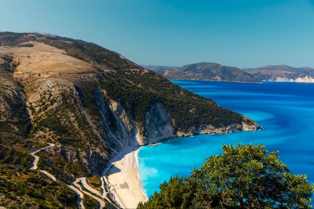 Aerial view of Myrtos Beach in Kefalonia, Greece, showing bright turquoise waters meeting a crescent-shaped white pebble shoreline backed by dramatic limestone cliffs and lush greenery. The winding road leading to the beach is visible on the left, emphasizing the remote and scenic setting.