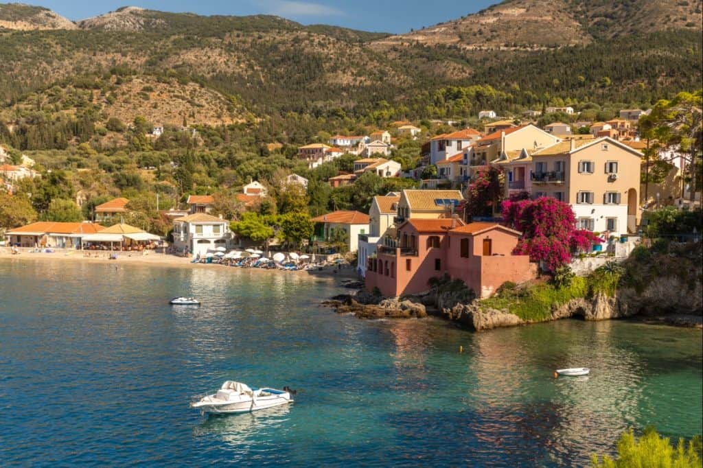 Coastal village of Assos in Kefalonia, Greece, featuring pastel-colored houses with terracotta roofs nestled along a calm, clear bay. Small boats float near the shore while vibrant bougainvillea adds a splash of color to the waterfront scene.87