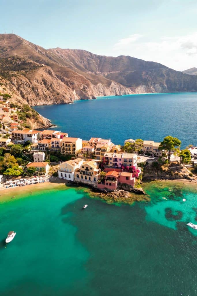 Aerial view of Assos village in Kefalonia, Greece, showing colorful seaside houses surrounded by turquoise waters and dramatic cliffs in the background. The peninsula layout and shallow emerald coves add to the village’s charm and secluded atmosphere.