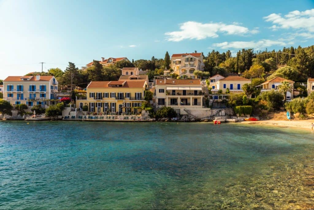 Waterfront view of Fiskardo village in Kefalonia, Greece, featuring colorful Venetian-style buildings with balconies and shuttered windows lining the calm, clear shoreline. The shallow turquoise waters meet a small sandy beach on the right, with lush greenery and hillside homes in the background.