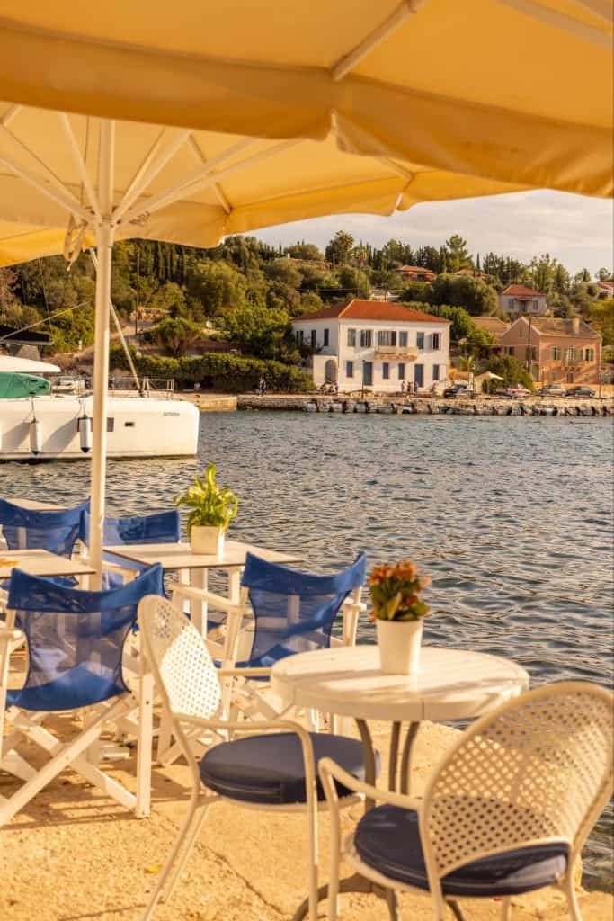 Outdoor café seating with white tables and blue chairs set under large cream umbrellas on the edge of Fiskardo’s harbor. The calm water reflects golden light and a white and blue building sits across the bay, framed by hillside greenery.