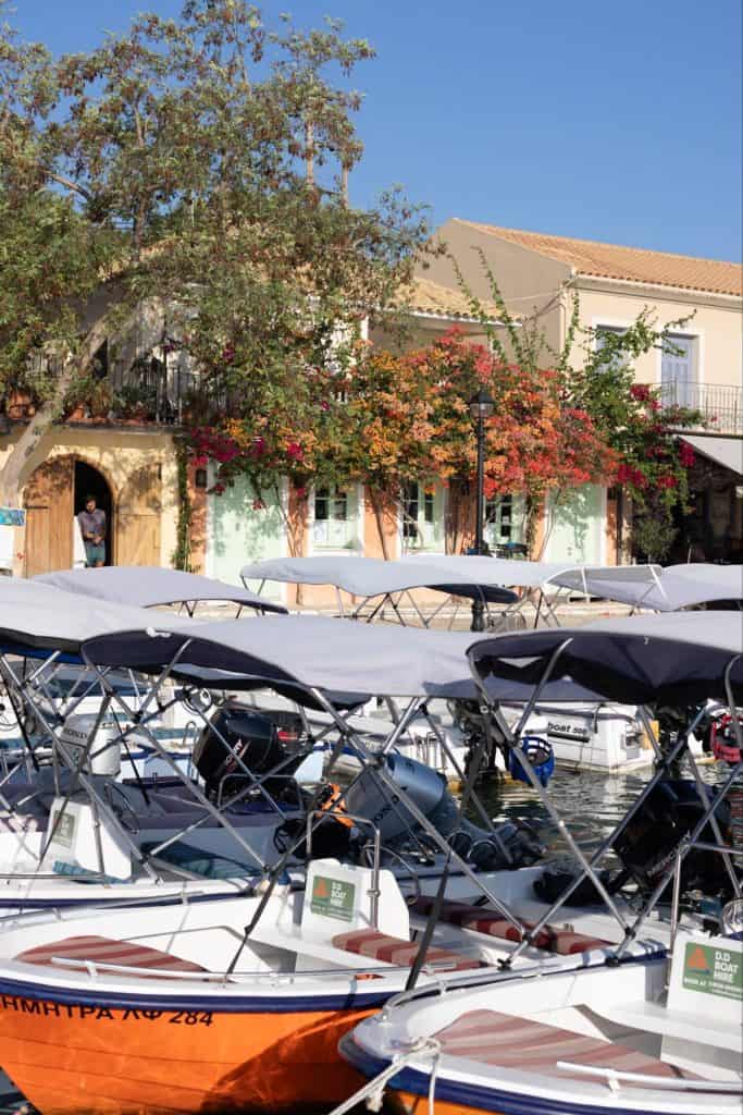 Docked boats with canvas canopies line the waterfront in Fiskardo, Kefalonia, with pastel-colored buildings and vibrant autumn vines visible in the background. A person stands in a doorway, adding a sense of daily village life to the serene harbor scene.