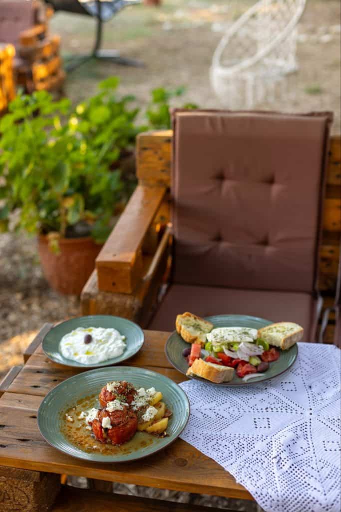 A rustic outdoor meal setting with three traditional Greek dishes: baked tomato stuffed with rice and topped with feta, a fresh village salad with bread and tzatziki, and a side of yogurt with a single olive. The food is served on a wooden table with a lace cloth beside a cushioned wooden chair.