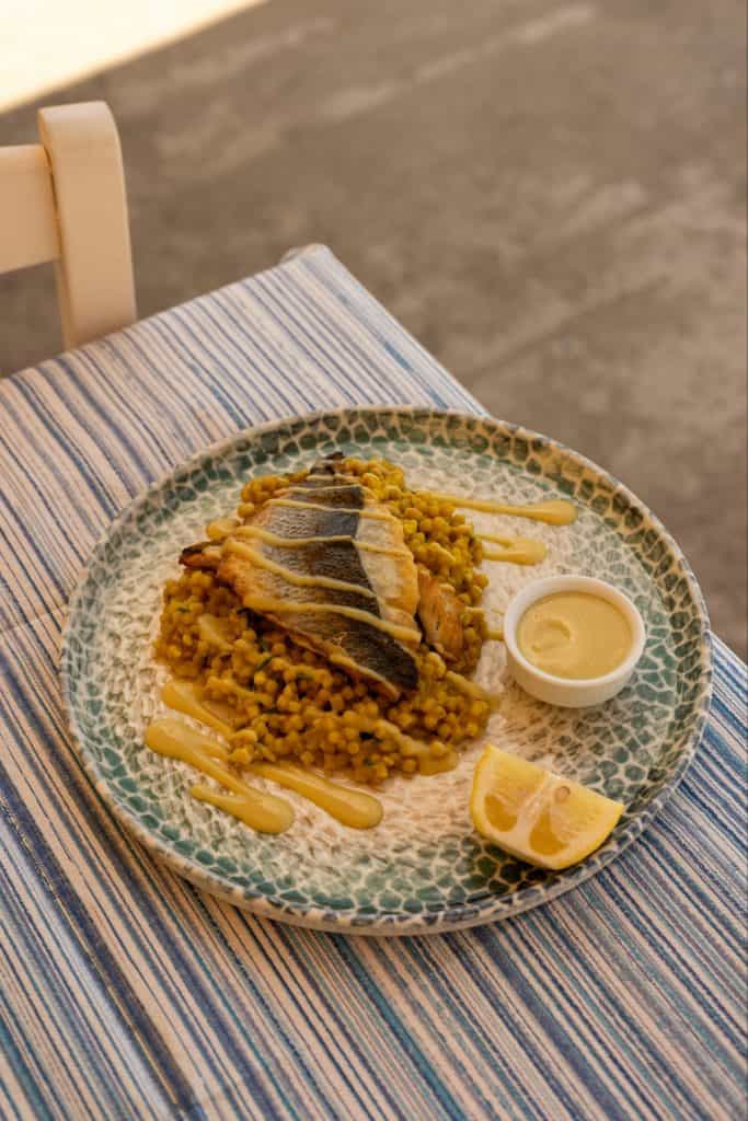 A Mediterranean seafood dish featuring grilled fish fillets served over a bed of pearl couscous with yellow spices, accompanied by a lemon wedge and creamy dipping sauce. The plate rests on a blue-striped tablecloth, enhancing the relaxed coastal dining vibe.