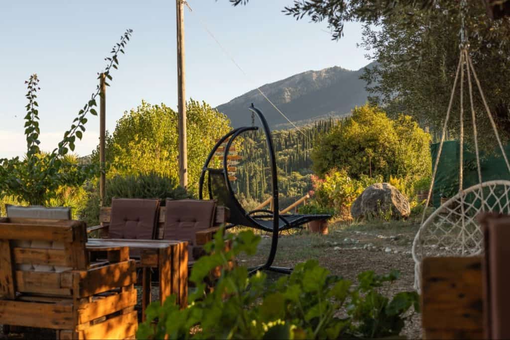 Rustic outdoor seating area in Kefalonia, Greece, featuring wooden pallet furniture, a hanging egg chair, and a rope hammock nestled among greenery. The peaceful garden overlooks a lush valley and forested mountain under soft golden hour light.