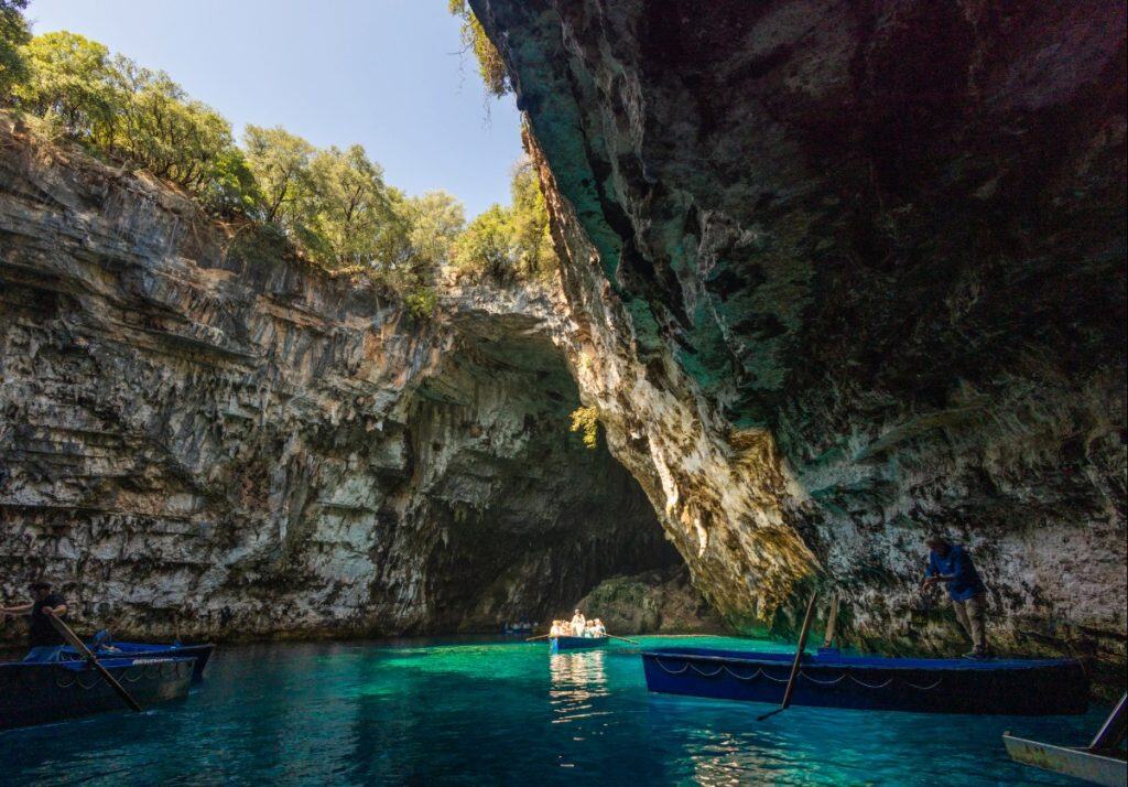 View from inside Melissani Cave in Kefalonia, Greece, showing sunlight pouring through a collapsed cave roof onto vivid turquoise water. Rowboats glide through the cave's crystal-clear lake, framed by dramatic limestone walls.