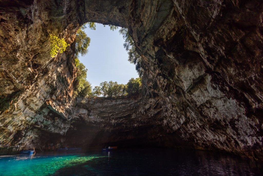 Wide-angle shot of Melissani Cave’s open ceiling, revealing the sky and trees above while sunlight beams down onto the deep blue water below. Small boats drift in the cave’s interior, highlighting its surreal, natural beauty.