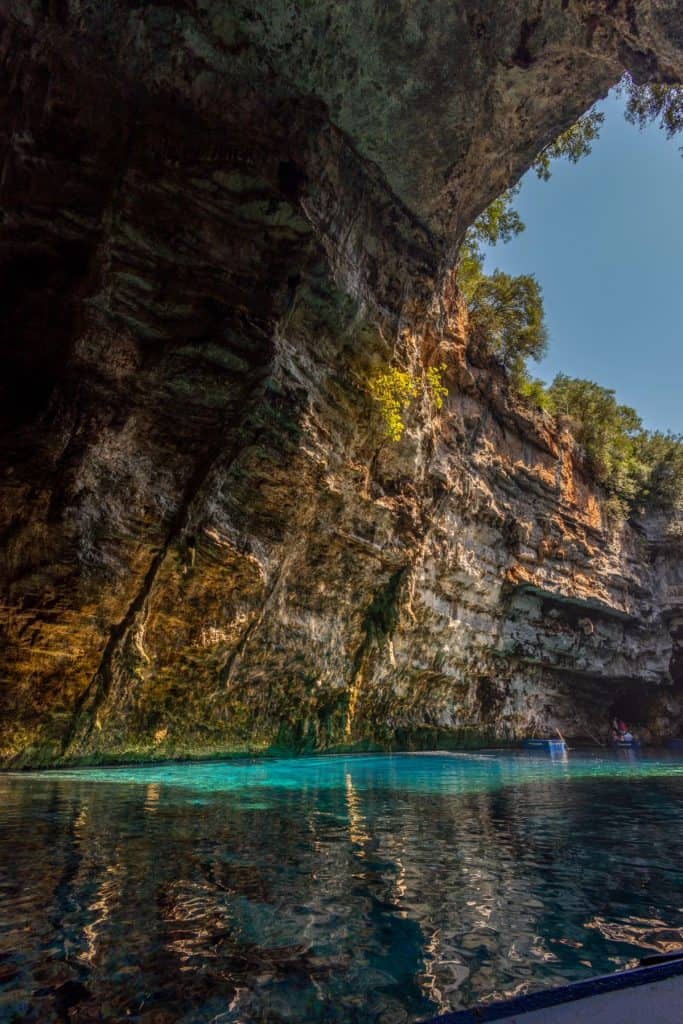 Interior of Melissani Cave showcasing the dramatic contrast between shadowed rock formations and the glowing aqua-blue lake below. Sunlight streams through the cave’s opening, illuminating patches of moss and greenery clinging to the stone walls.